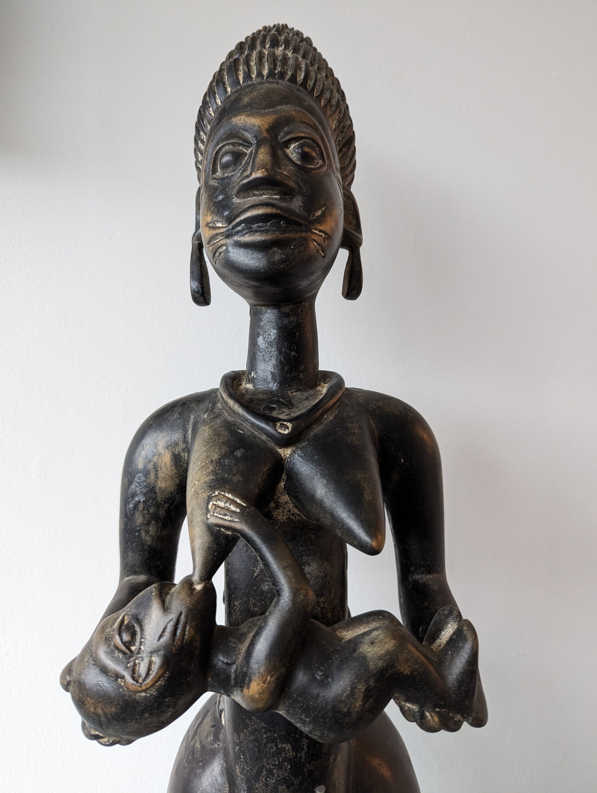 A front, close-up view of a dark wooden Yoruba figural statue of a woman breastfeeding a baby. The statue has intricate carvings on the woman's hair, face, and body.
