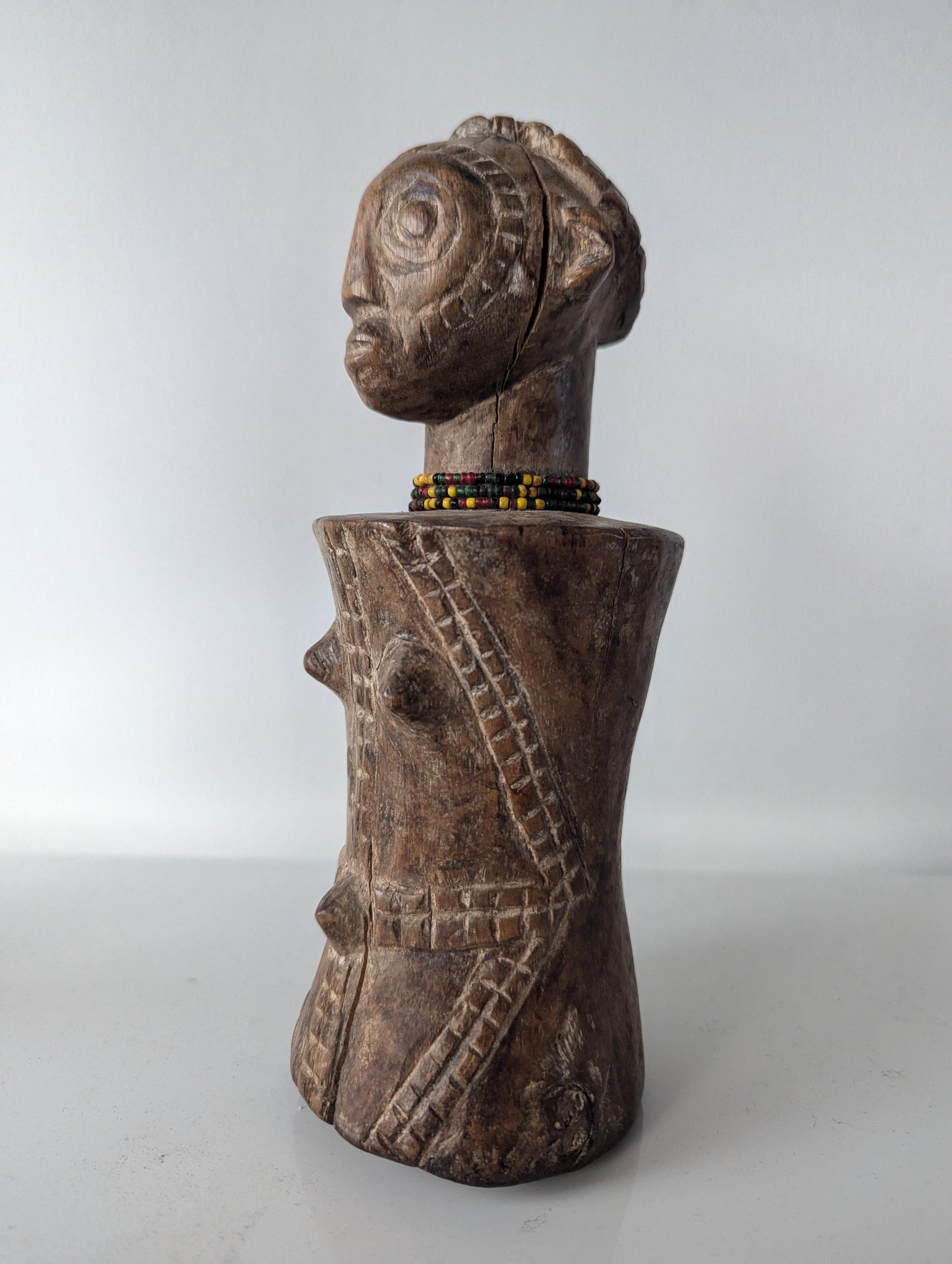 A close-up, frontal view of a dark, wooden Tabwa Mpundu doll. The figure has a distinctive head with detailed carving, including hair and facial features. A small beaded necklace, with yellow and black beads, sits at the base of its neck. The torso is bulbous and rounded, with carved tribal patterns and two protruding points on the chest.