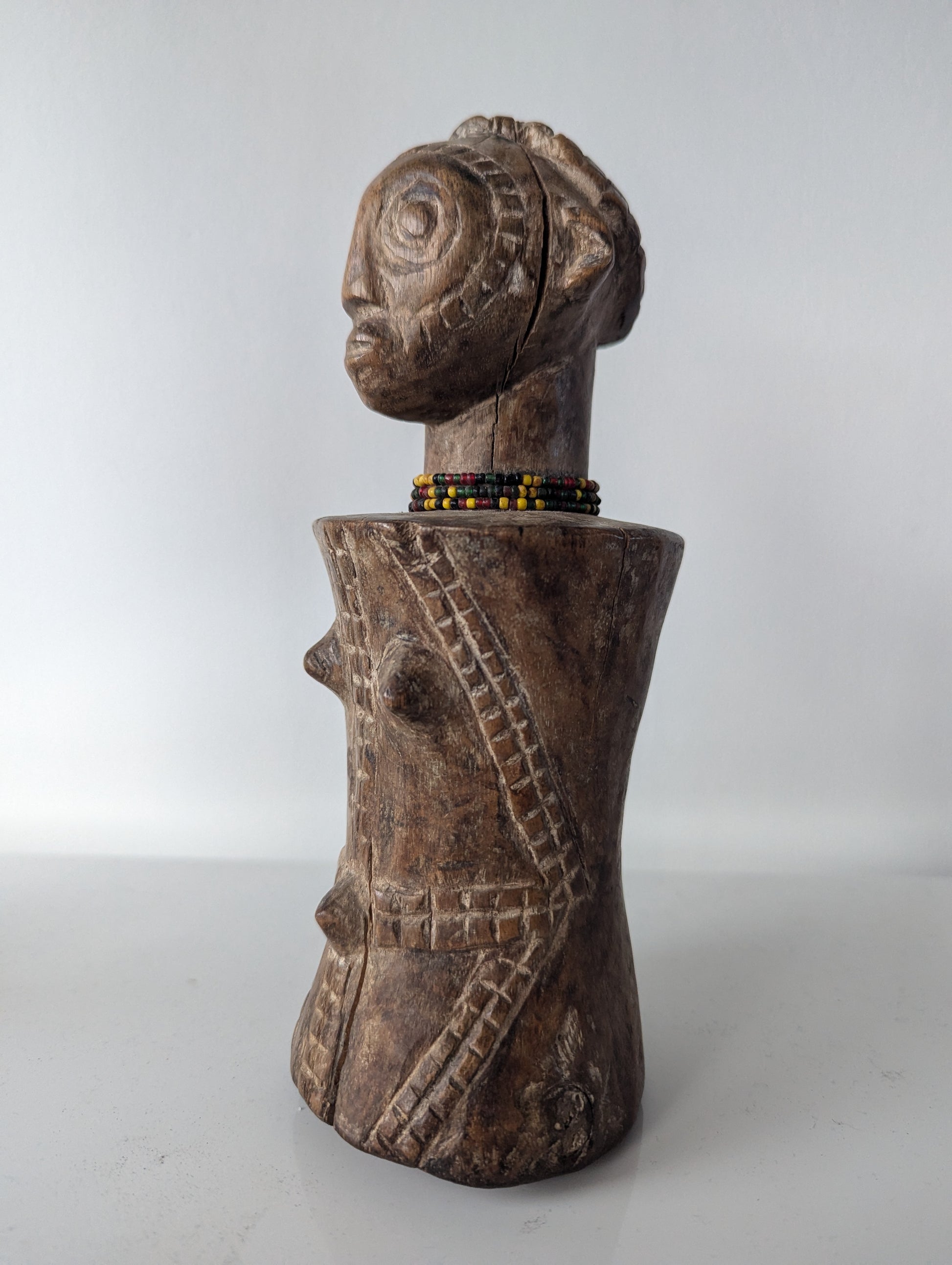 A close-up, frontal view of a dark, wooden Tabwa Mpundu doll. The figure has a distinctive head with detailed carving, including hair and facial features. A small beaded necklace, with yellow and black beads, sits at the base of its neck. The torso is bulbous and rounded, with carved tribal patterns and two protruding points on the chest.