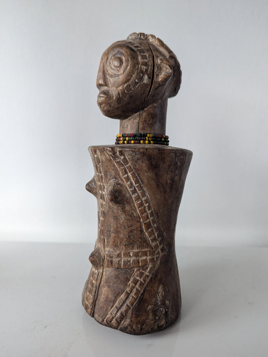 A close-up, frontal view of a dark, wooden Tabwa Mpundu doll. The figure has a distinctive head with detailed carving, including hair and facial features. A small beaded necklace, with yellow and black beads, sits at the base of its neck. The torso is bulbous and rounded, with carved tribal patterns and two protruding points on the chest.