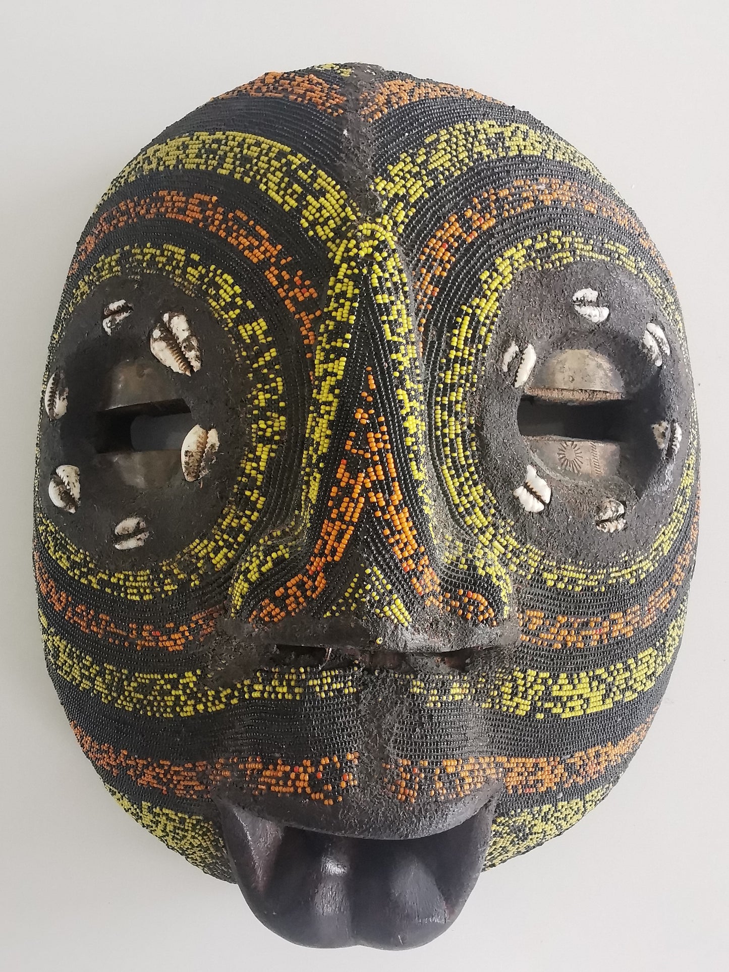A wooden mask with brass inlay eyes, decorated with colored beads and cowry shells, featuring a guppy-shaped mouth.