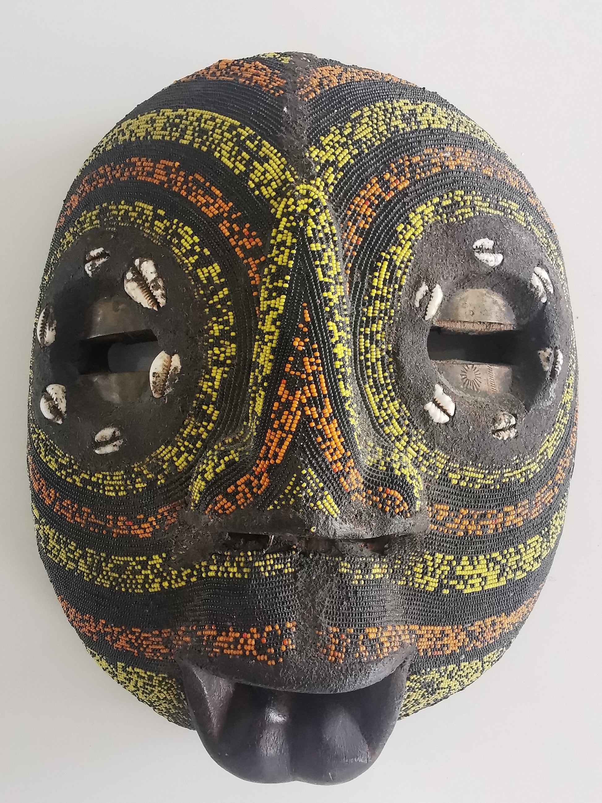 A wooden mask with brass inlay eyes, decorated with colored beads and cowry shells, featuring a guppy-shaped mouth.