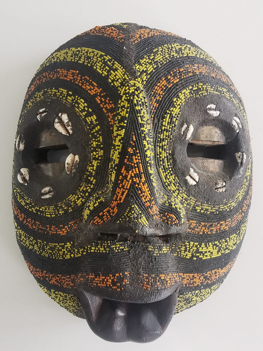A wooden mask with brass inlay eyes, decorated with colored beads and cowry shells, featuring a guppy-shaped mouth.
