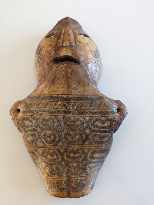 A carved wooden Atoni amulet, featuring a stylized human face at the top and intricate geometric and spiral patterns below. The piece has a light brown, aged texture with darker spots and is shaped like a flattened, elongated shield.