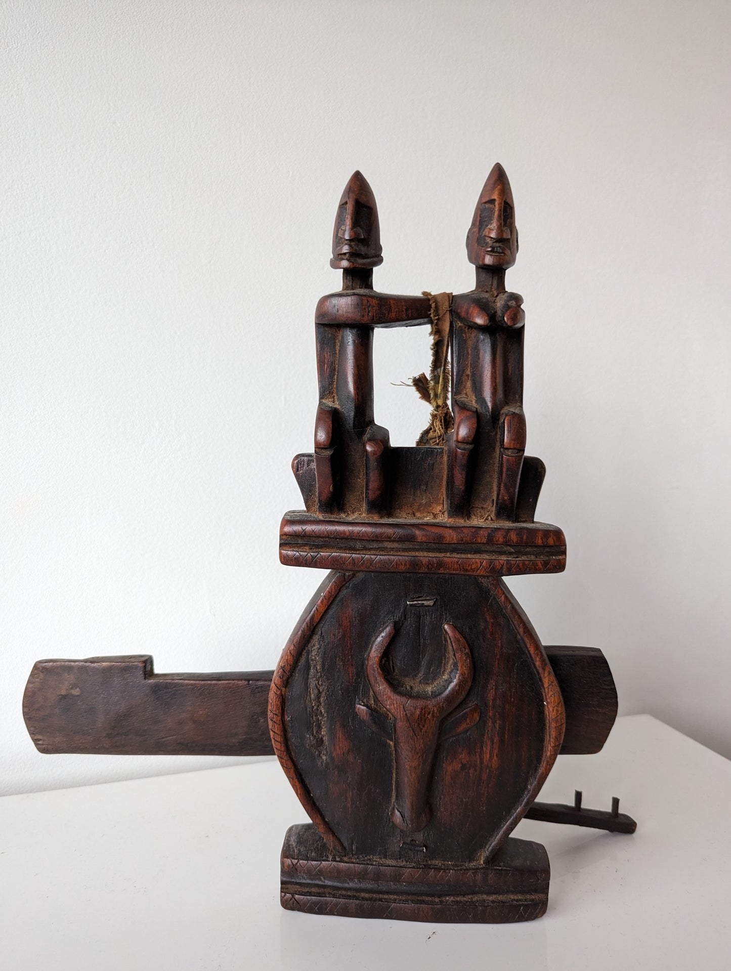 A full front view of a Dogon carved wooden granary door lock with a key. The lock features a prominent carved couple at the top and a circular, stylized face at the center.