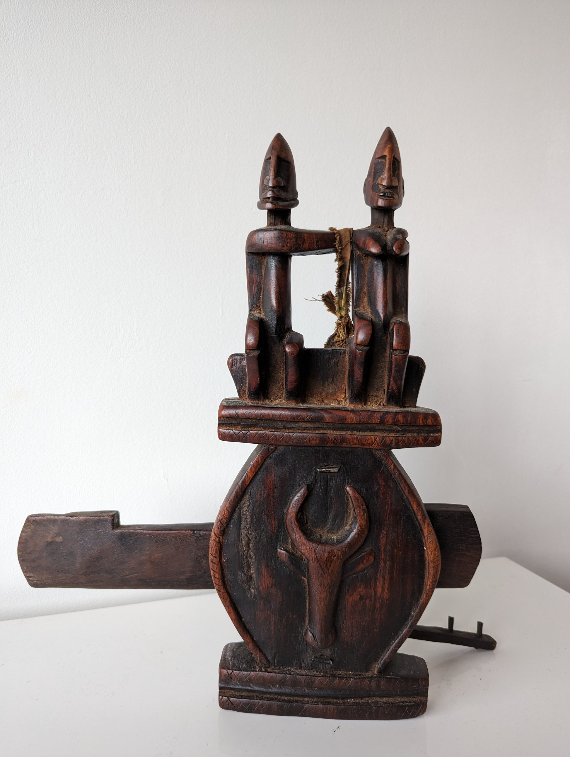 A full front view of a Dogon carved wooden granary door lock with a key. The lock features a prominent carved couple at the top and a circular, stylized face at the center.