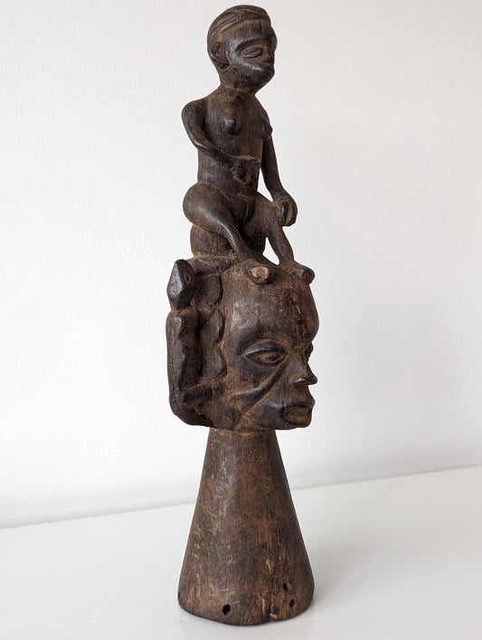 A wooden sculpture of an Ekoi headdress featuring a female face with a child on top and two animal figures on each side, with drilled apertures for hanging.