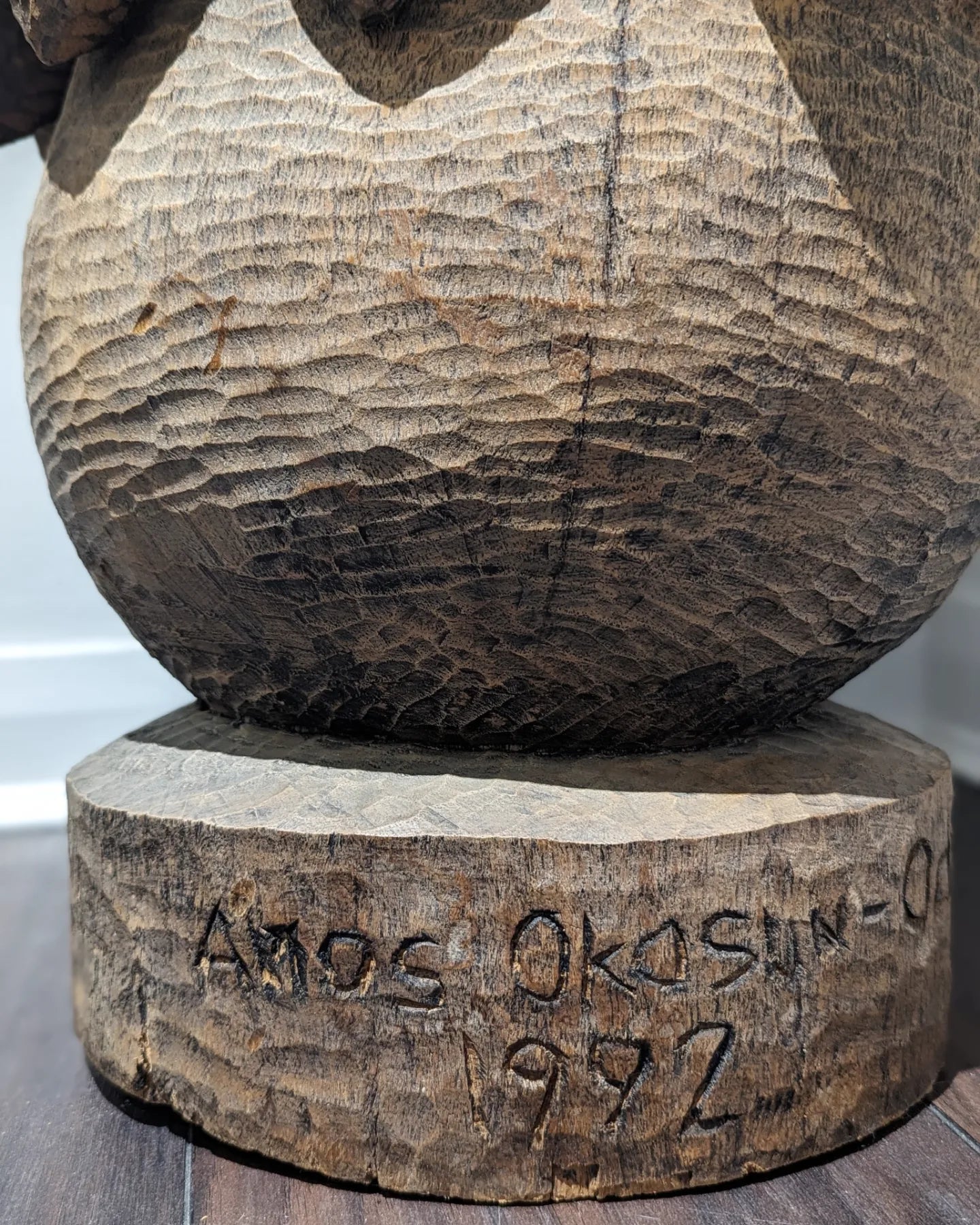 A close-up of the textured, circular base of the sculpture, with a visible artist's signature Amos Okosun Odion carved into the wood.