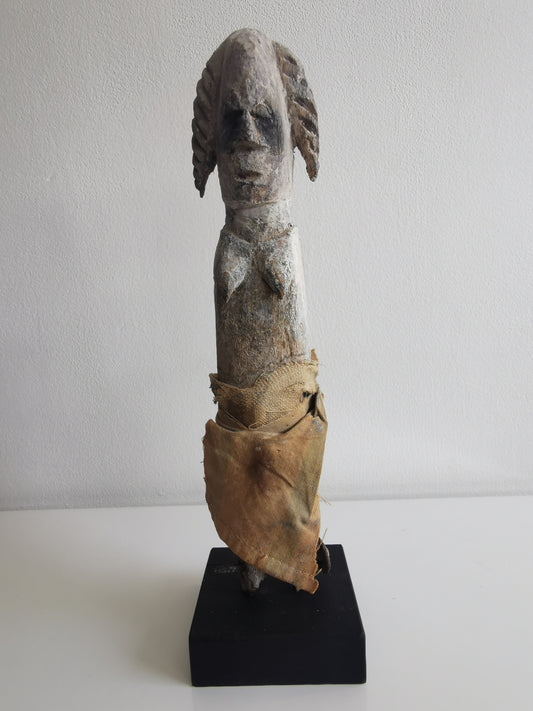 A front view of an Ibibio female statuette on a black display stand. The anthropomorphic statue is made of carved wood, has a prominent head and elongated neck, and is partially wrapped in cloth.