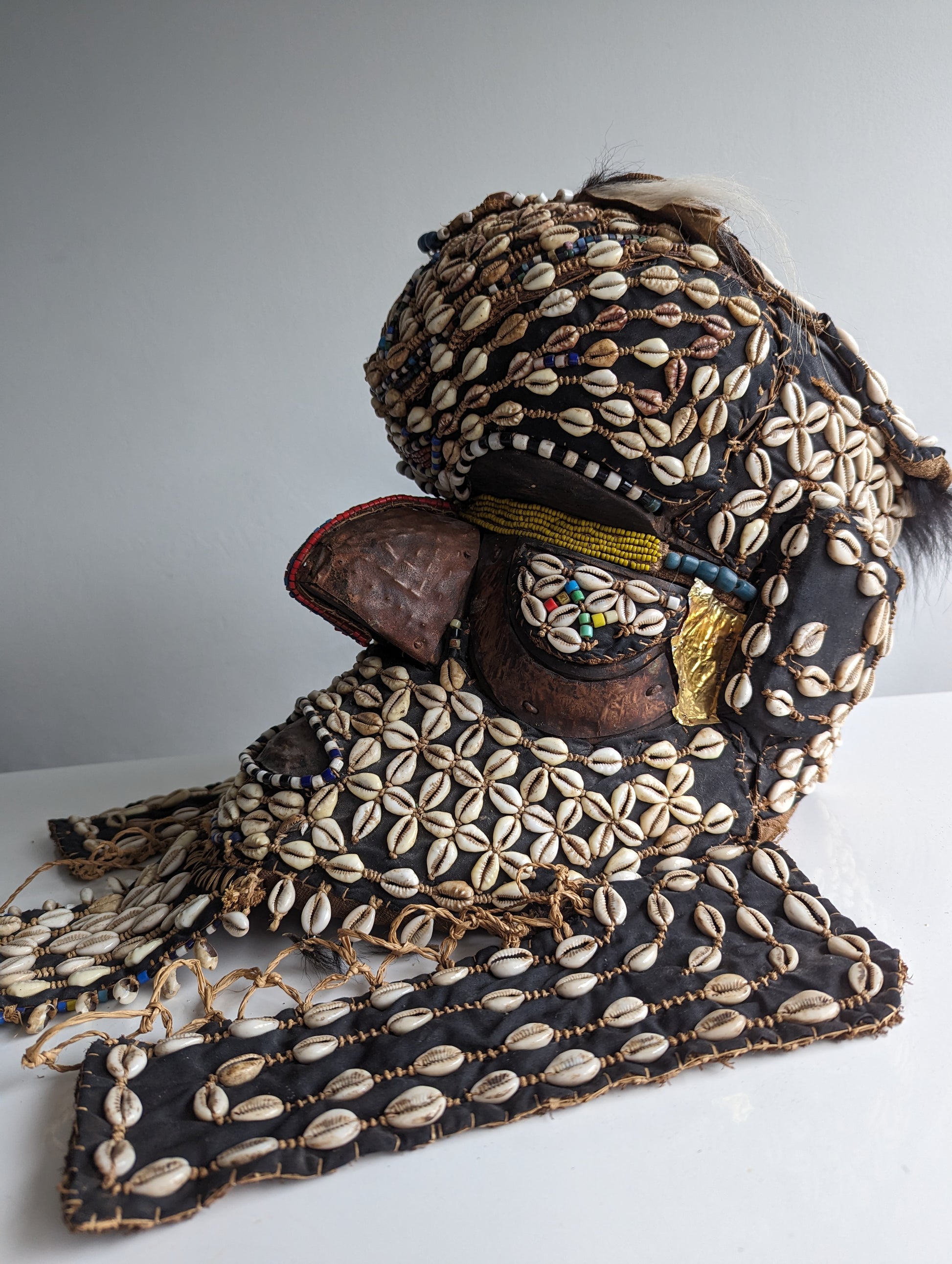 A three-quarter view of a ceremonial Kuba 'Bwoom' helmet mask from Congo. The mask is richly decorated with cowrie shells, beads, and fabric, with a prominent forehead and face.