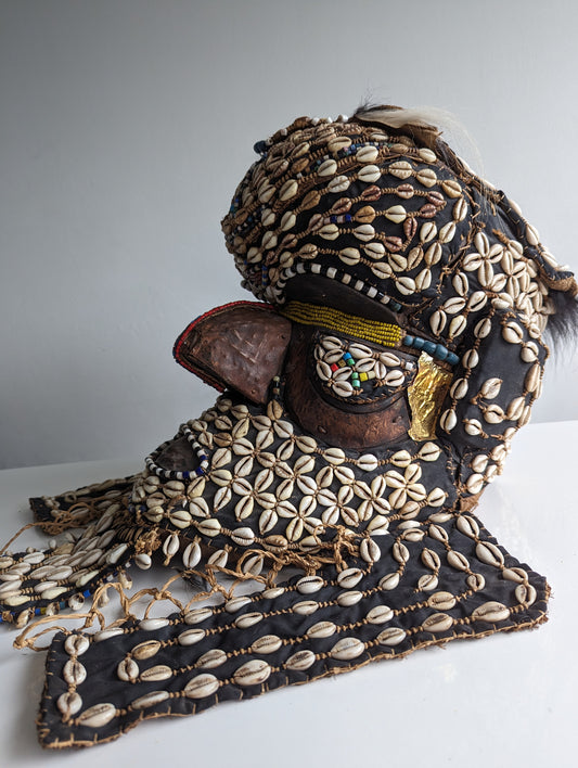 A three-quarter view of a ceremonial Kuba 'Bwoom' helmet mask from Congo. The mask is richly decorated with cowrie shells, beads, and fabric, with a prominent forehead and face.