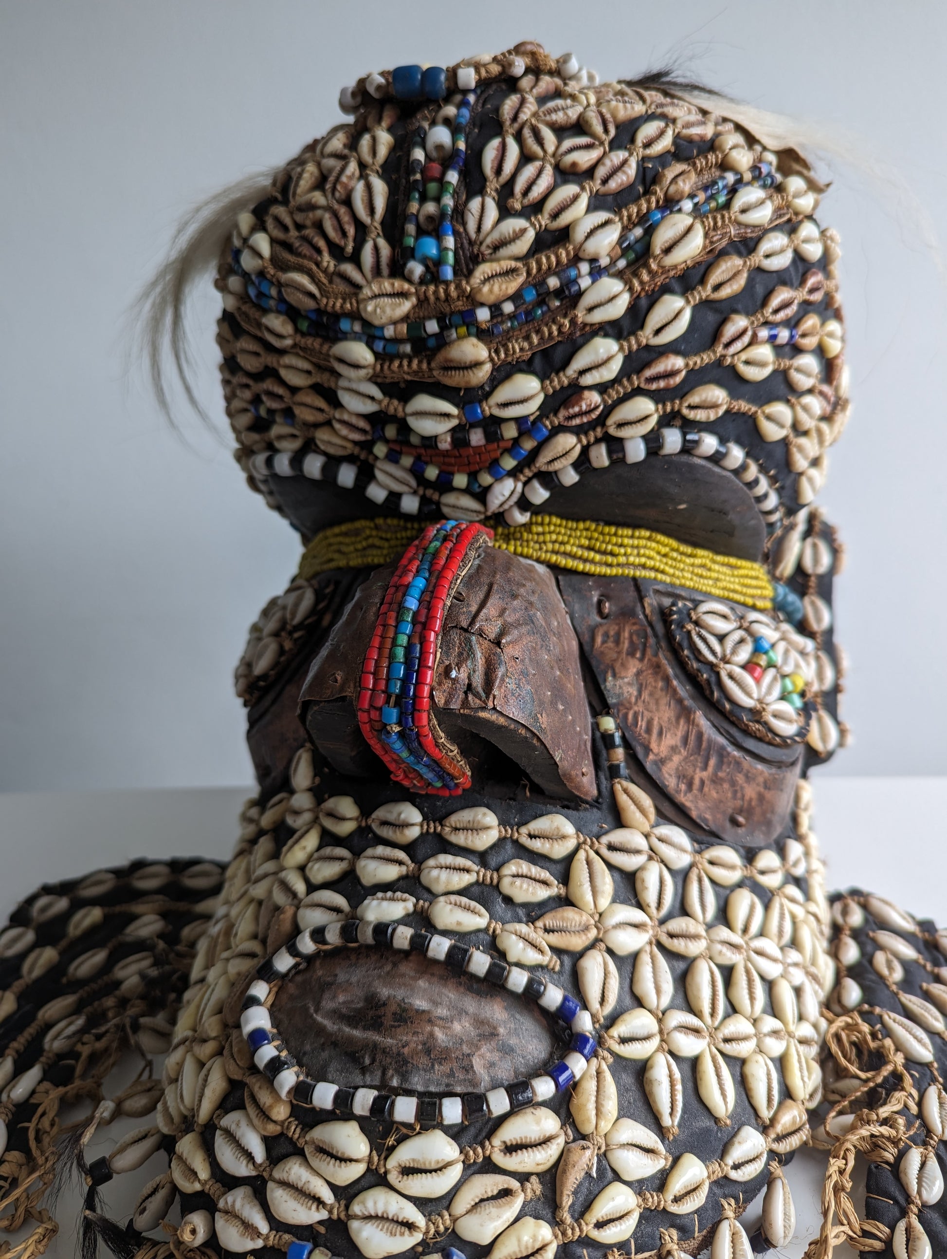 An angled, close-up view of the mask's face and upper torso, highlighting the intricate patterns made from cowrie shells and the details of the beaded collar