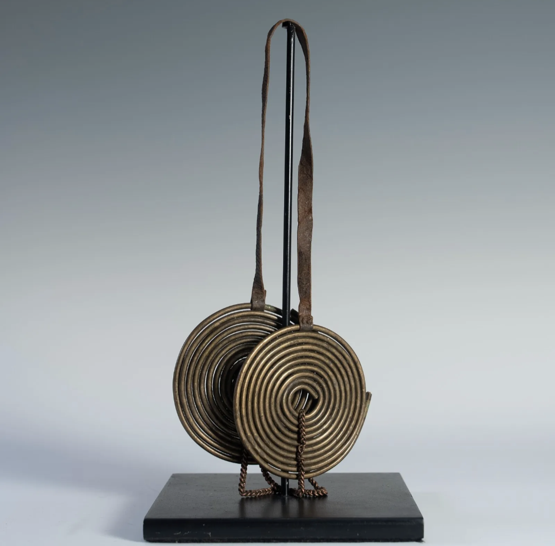 A detailed view of a Maasai coiled brass neck adornment, called 'Taet Tai,' on a black display stand. The piece consists of two large, circular, coiled brass disks suspended from a curved brass support.