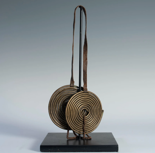 A detailed view of a Maasai coiled brass neck adornment, called 'Taet Tai,' on a black display stand. The piece consists of two large, circular, coiled brass disks suspended from a curved brass support.