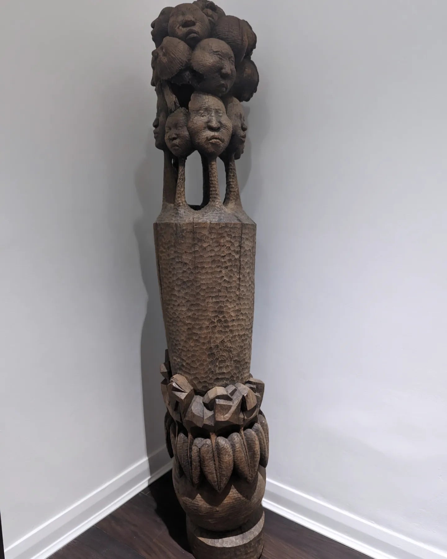A tall, full view of a 'Tree of Life' sculpture by Nigerian artist Amos Okosun Odion. The carved wooden piece features a series of human faces stacked on top of a textured column, with a wide, patterned base