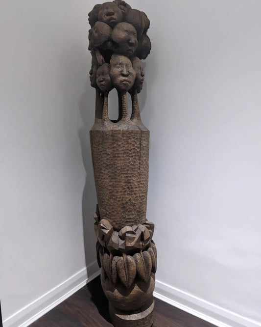 A tall, full view of a 'Tree of Life' sculpture by Nigerian artist Amos Okosun Odion. The carved wooden piece features a series of human faces stacked on top of a textured column, with a wide, patterned base