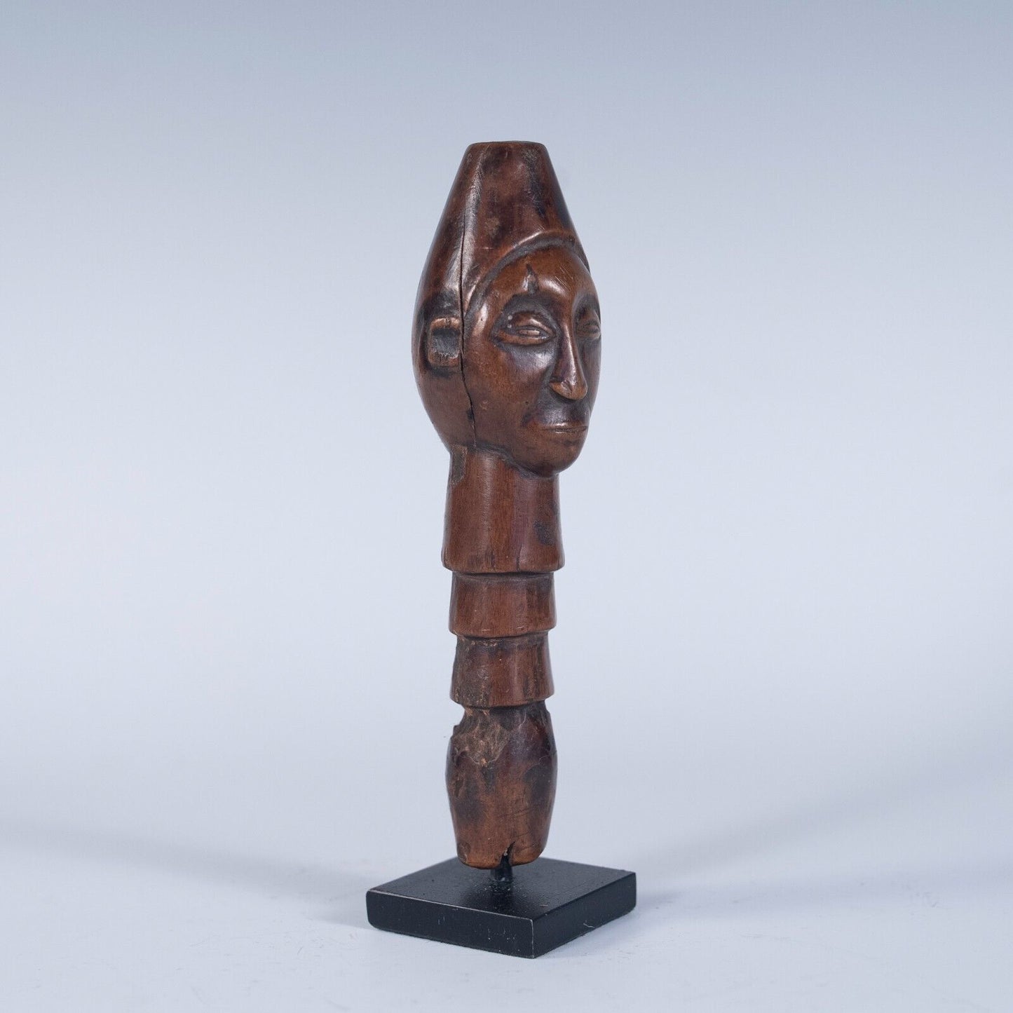 A front, slightly angled view of a tribal wooden top of a staff handle, featuring a carved human face. The handle has a segmented body and is displayed on a small black stand.