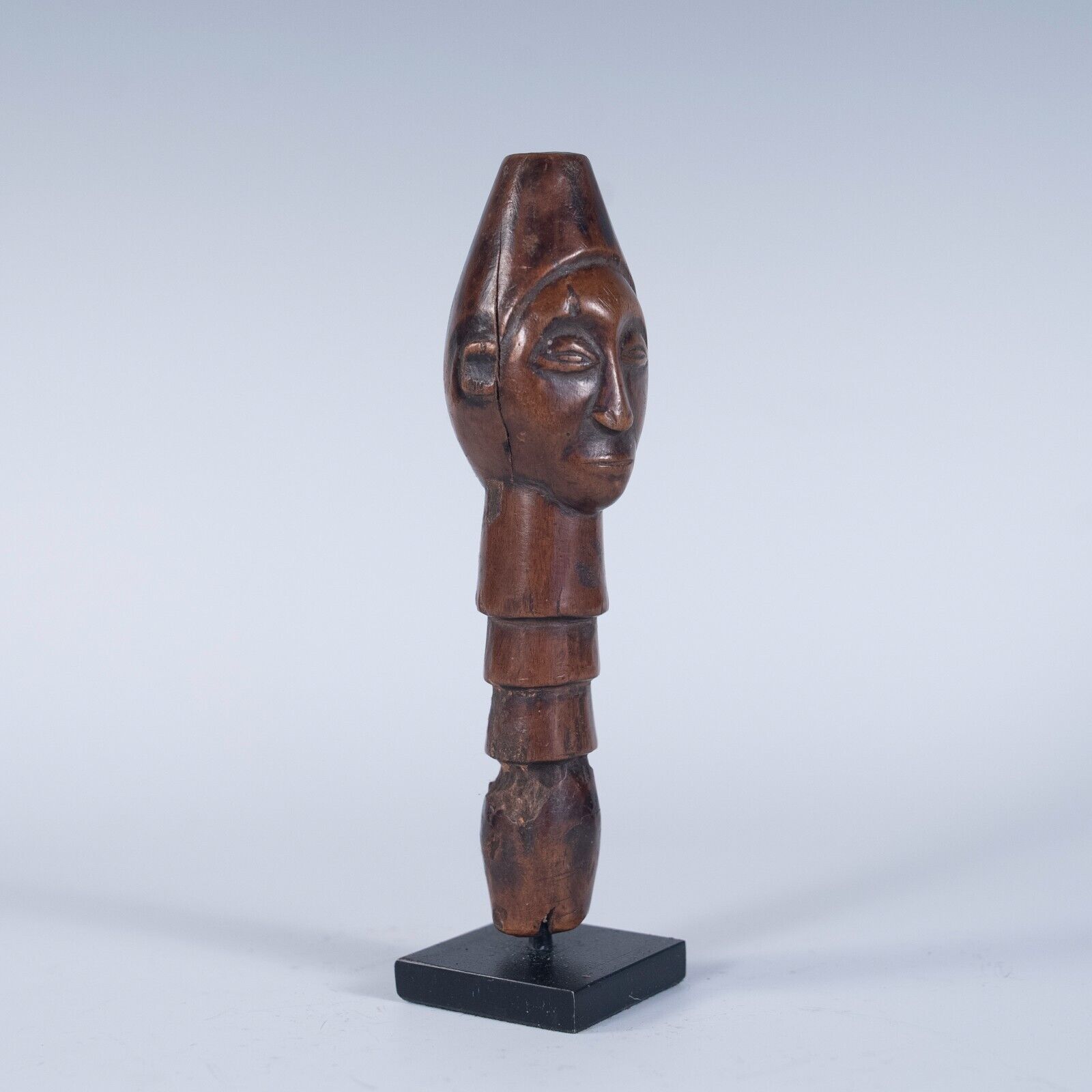 A front, slightly angled view of a tribal wooden top of a staff handle, featuring a carved human face. The handle has a segmented body and is displayed on a small black stand.