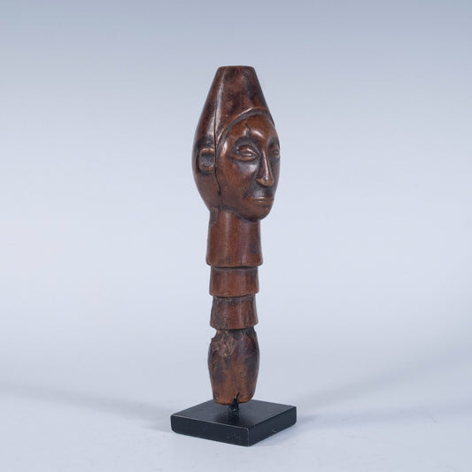 A front, slightly angled view of a tribal wooden top of a staff handle, featuring a carved human face. The handle has a segmented body and is displayed on a small black stand.