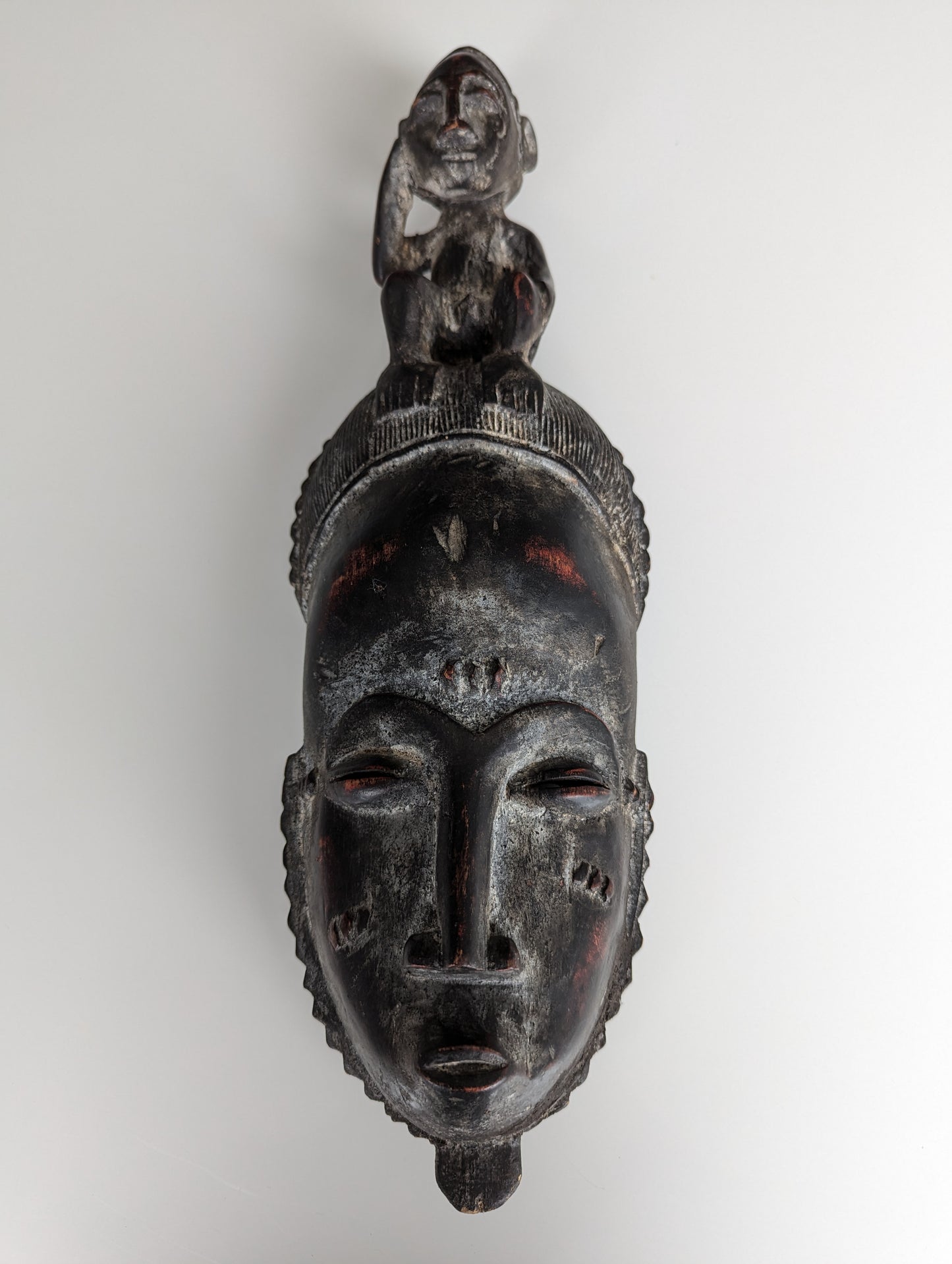 A front view of a Yaoure Baule (Baoulé) mask. The dark, carved wooden mask features an elongated face with slit eyes, a small mouth, and scarification marks, topped with a seated human figure.
