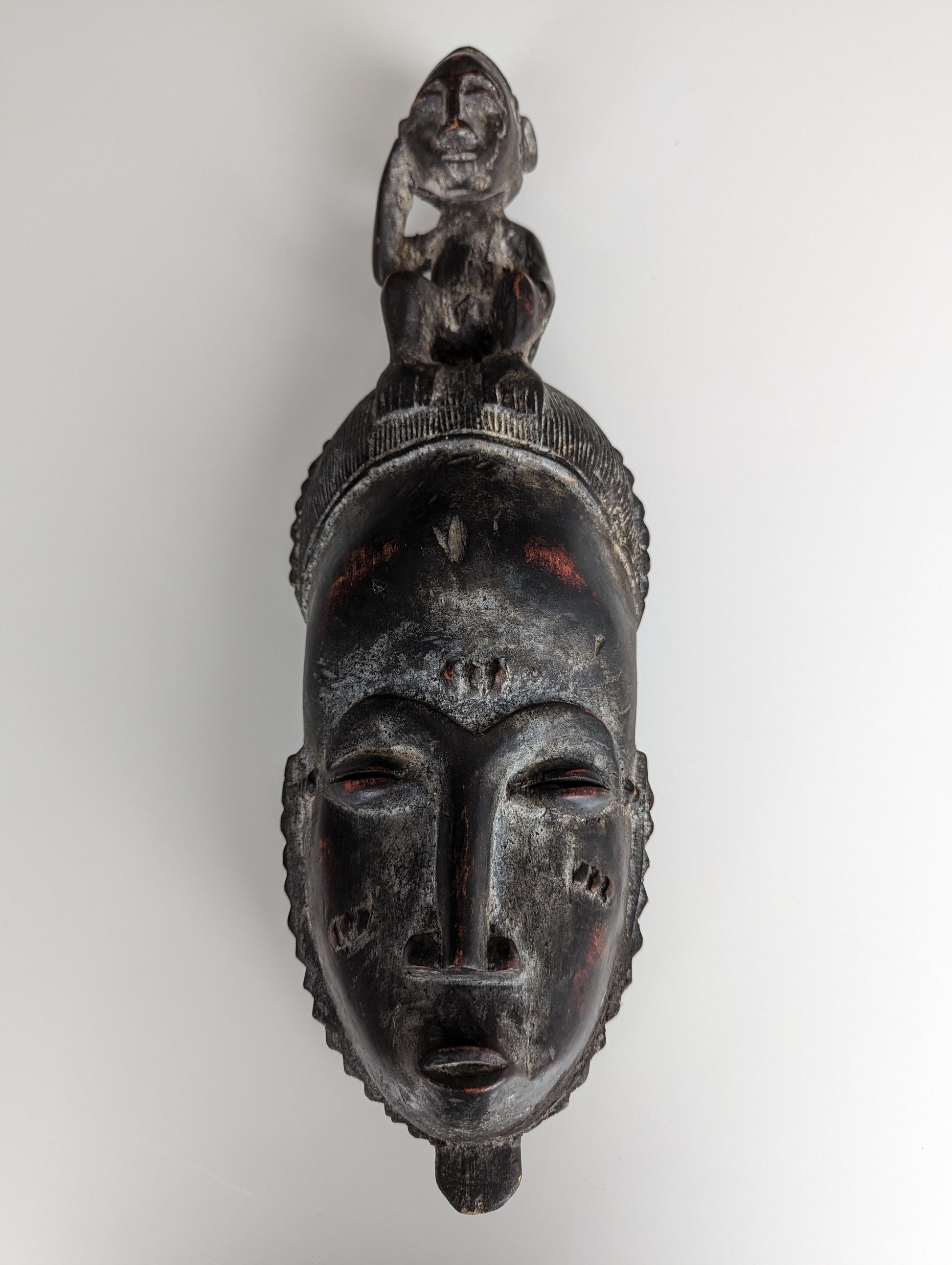 A front view of a Yaoure Baule (Baoulé) mask. The dark, carved wooden mask features an elongated face with slit eyes, a small mouth, and scarification marks, topped with a seated human figure.