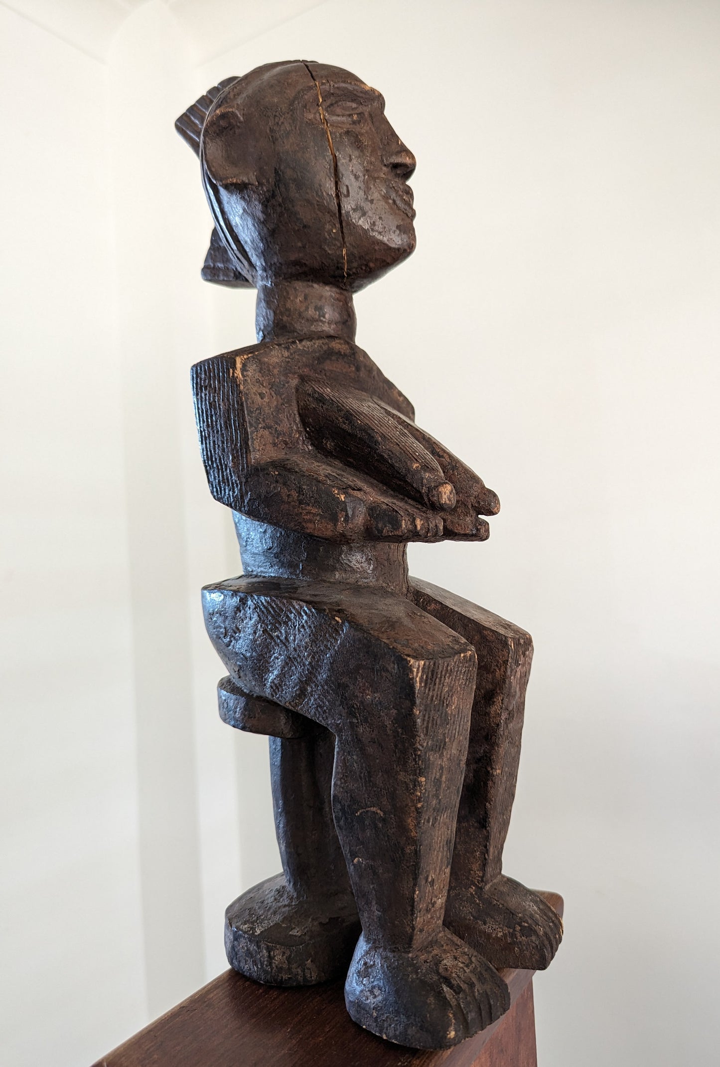 A front, three-quarter view of a Yoruba carved wooden statue of a seated female figure. The figure has a long neck, an angular face