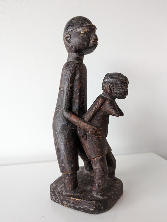 A wooden Lobi sculpture of two figures standing in an embrace, one behind the other. The figures have a dark, smooth finish and stylized features.