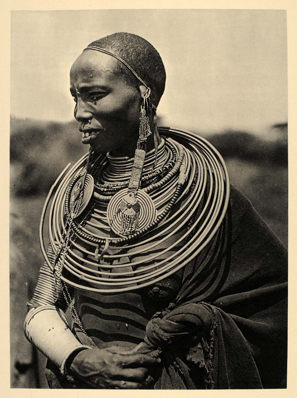 A historical black-and-white photograph of a Maasai woman wearing multiple brass and beaded neck adornments, similar to the one being sold