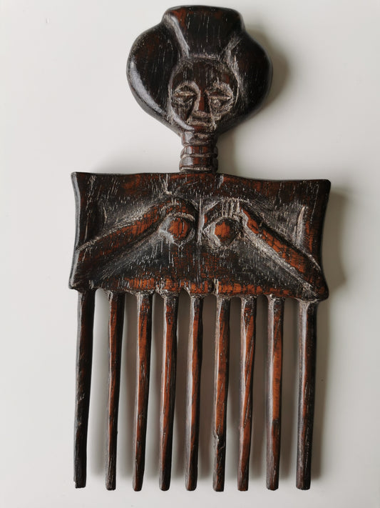 A full front view of an African Ashanti female wood hair comb. The comb is carved with a stylized female figure, featuring a prominent face, long neck, and a torso with hands resting below the breasts, which form the handle above the teeth of the comb.