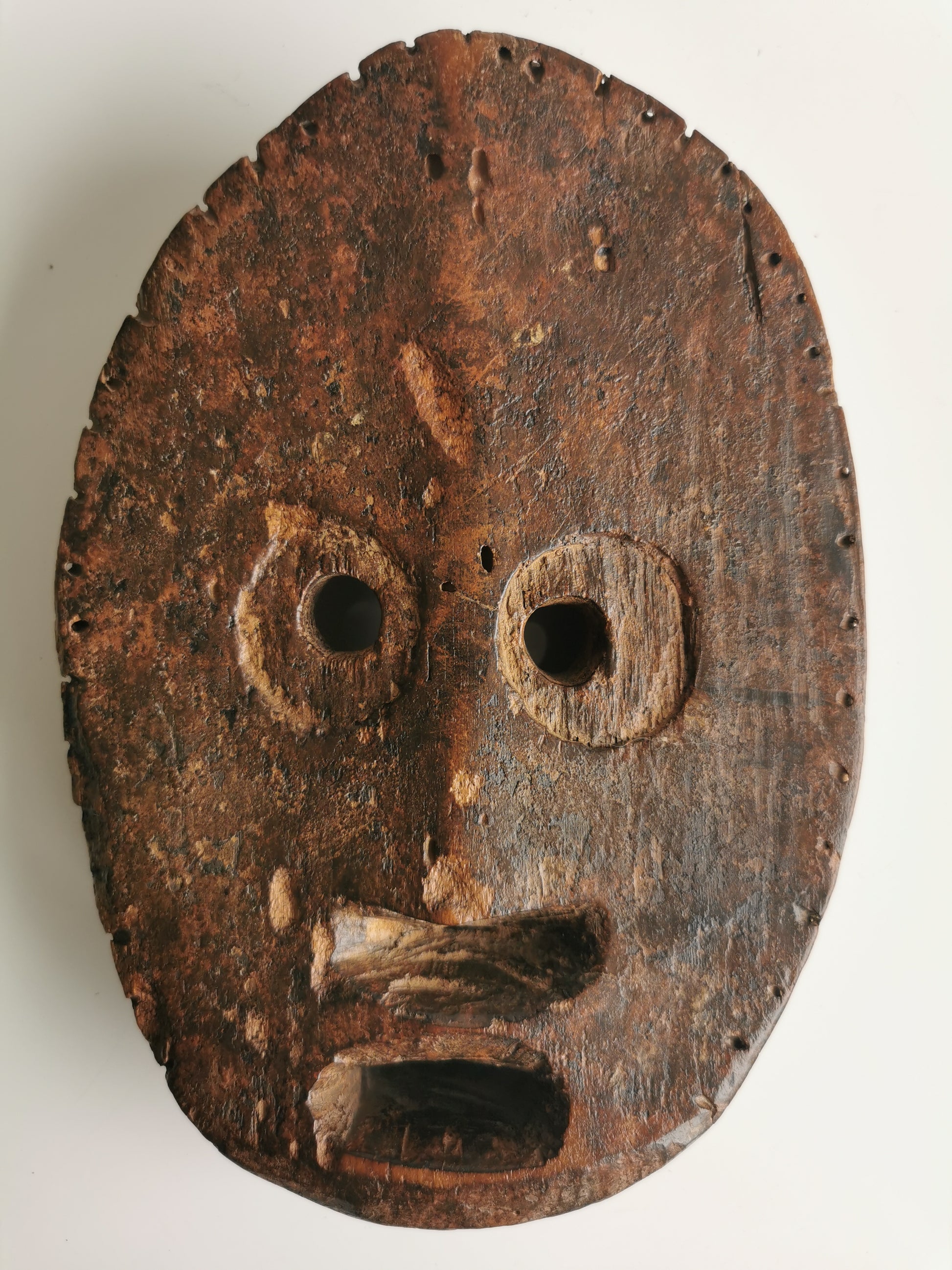 The image depicts a circular, wooden mask with a dark brown patina. The mask has two large, circular eye holes and a wide, open mouth. The facial features are carved in a simple, stylized manner. The mask appears to be quite old and shows signs of wear and tear.