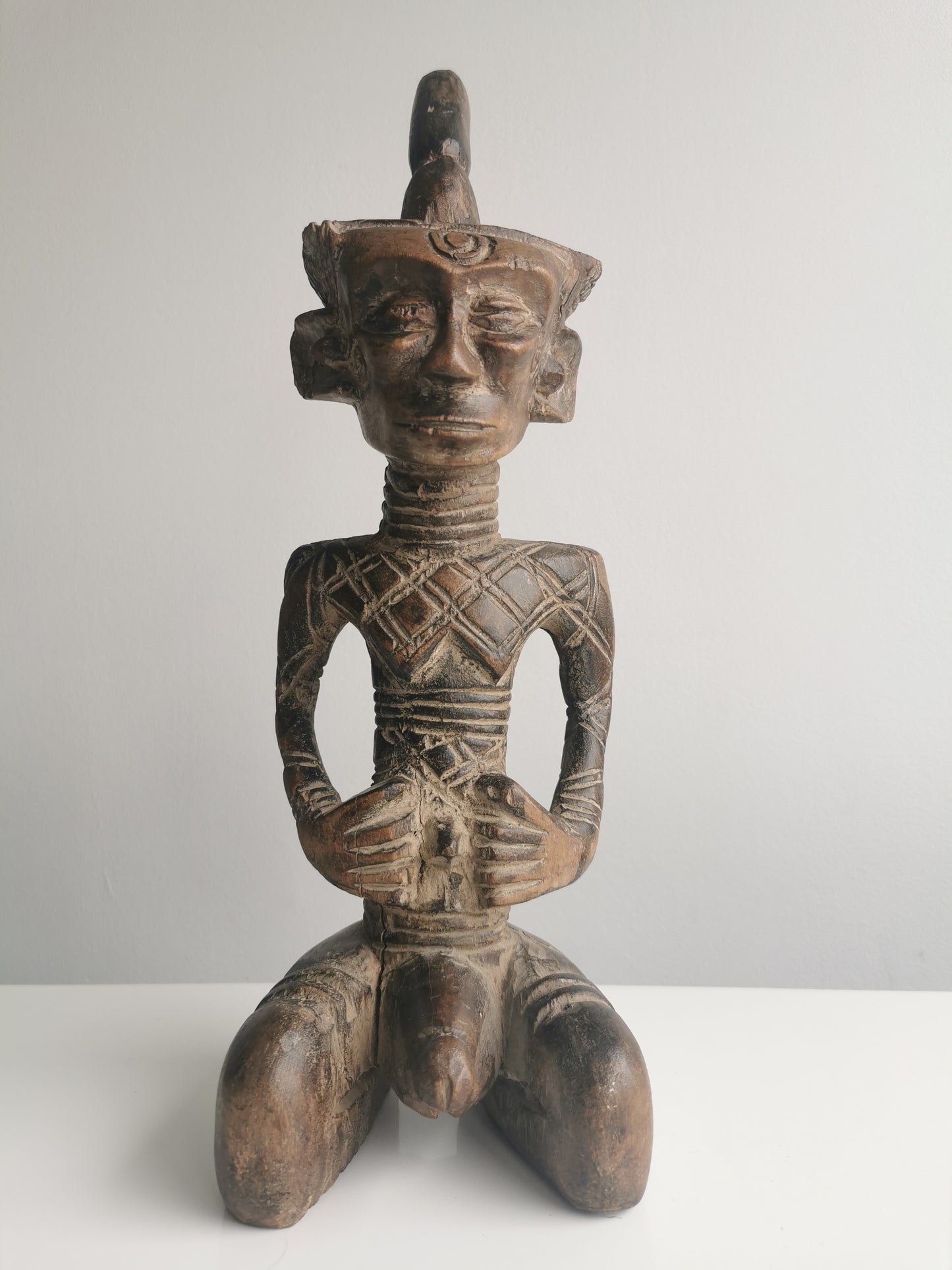 A front view of a Dengese carved wooden statue of a seated male figure. The statue features an elongated head with a tall headdress and a torso with incised geometric patterns.