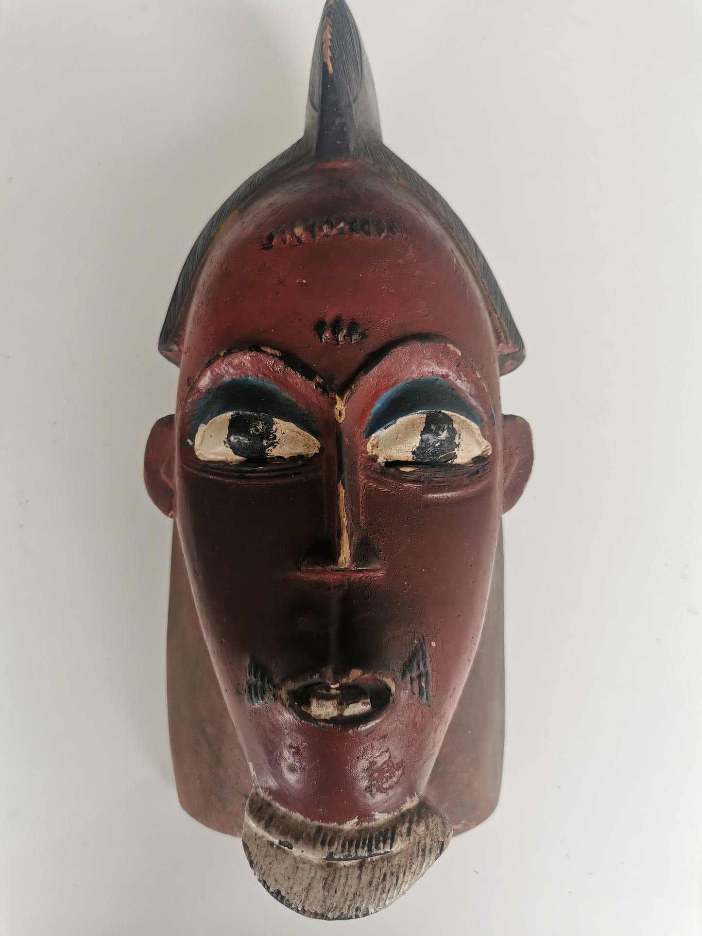 A front view of a Mid-Century red Guro 'Yu Gu' mask. The carved wooden mask has an elongated face, a prominent nose, and a tall, stylized headdress, with a vibrant red and black patina.