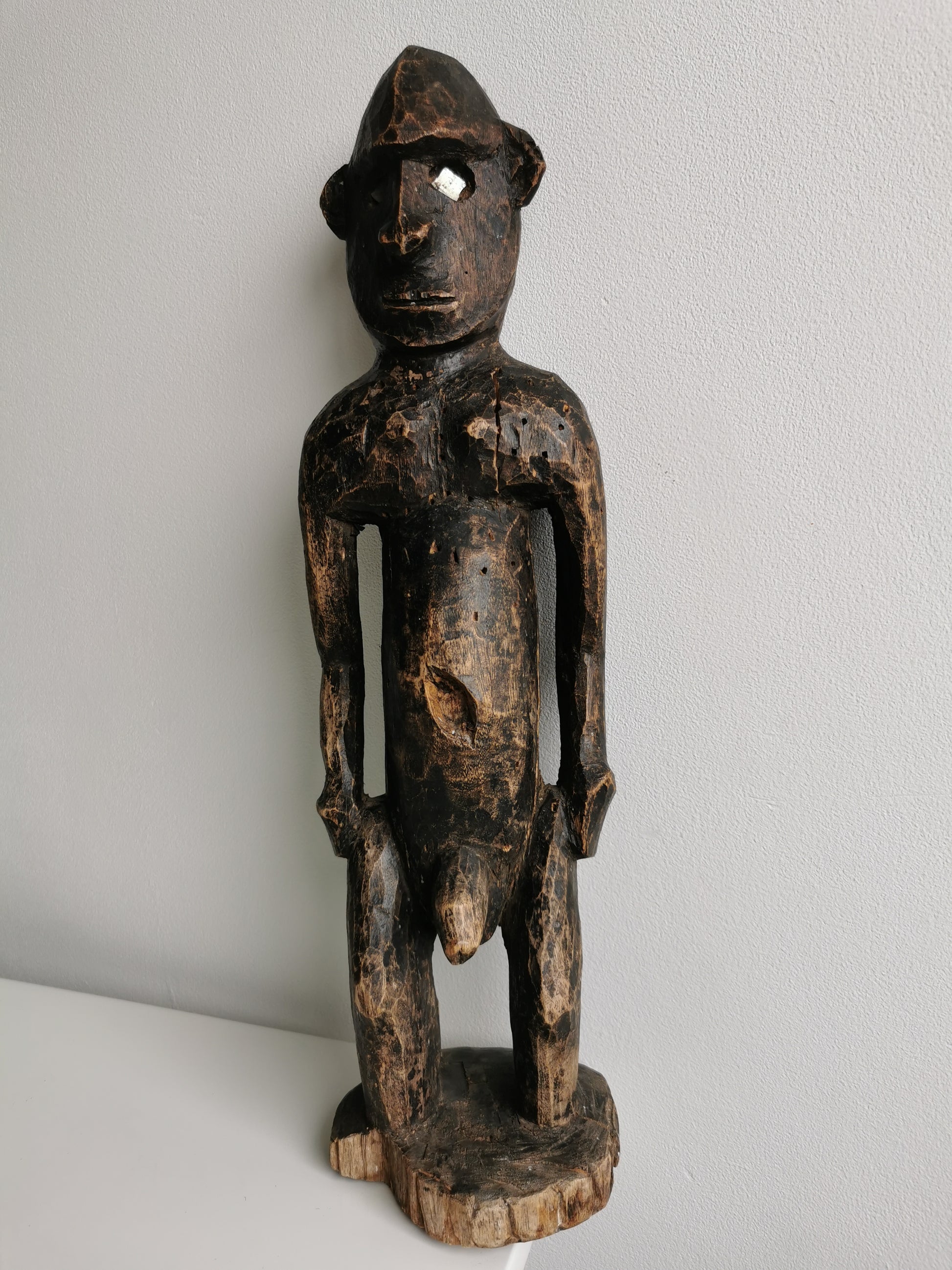 A front view of a West African male ancestor statue carved from dark, aged wood. The statue has a prominent head, a slim body, and light-colored material in the eyes.