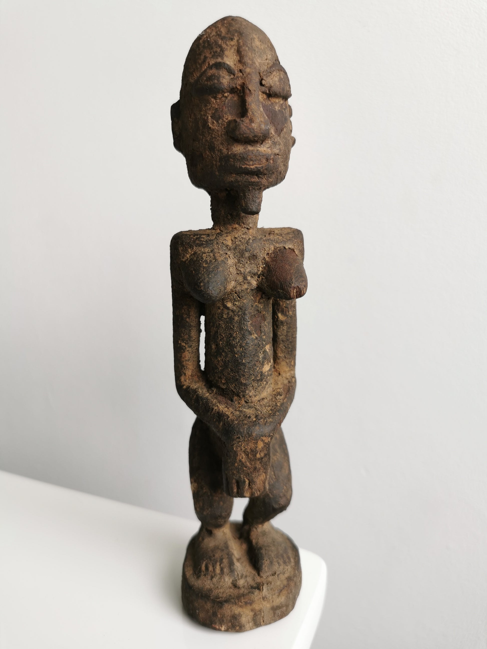 Frontal view of a dark, aged wooden Lobi tribal female ancestor figure statuette. The figure has a rounded head with carved facial features, including closed eyes and a small mouth. Its arms are positioned with hands resting over its abdomen, and it stands with slightly bent knees on a small, circular base.