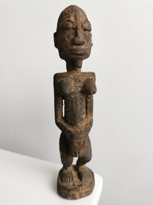 Frontal view of a dark, aged wooden Lobi tribal female ancestor figure statuette. The figure has a rounded head with carved facial features, including closed eyes and a small mouth. Its arms are positioned with hands resting over its abdomen, and it stands with slightly bent knees on a small, circular base.